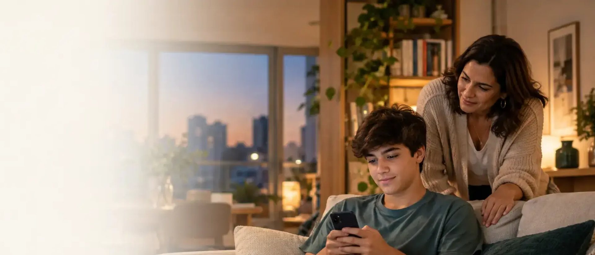 Adolescente sentado no sofá olhando o celular ao entardecer enquanto uma mulher se aproxima por trás, com expressão calma, para conversar